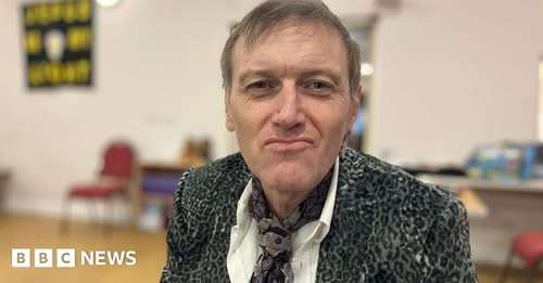 Man in a patterned blazer with a serious expression, in a room with chairs and books.